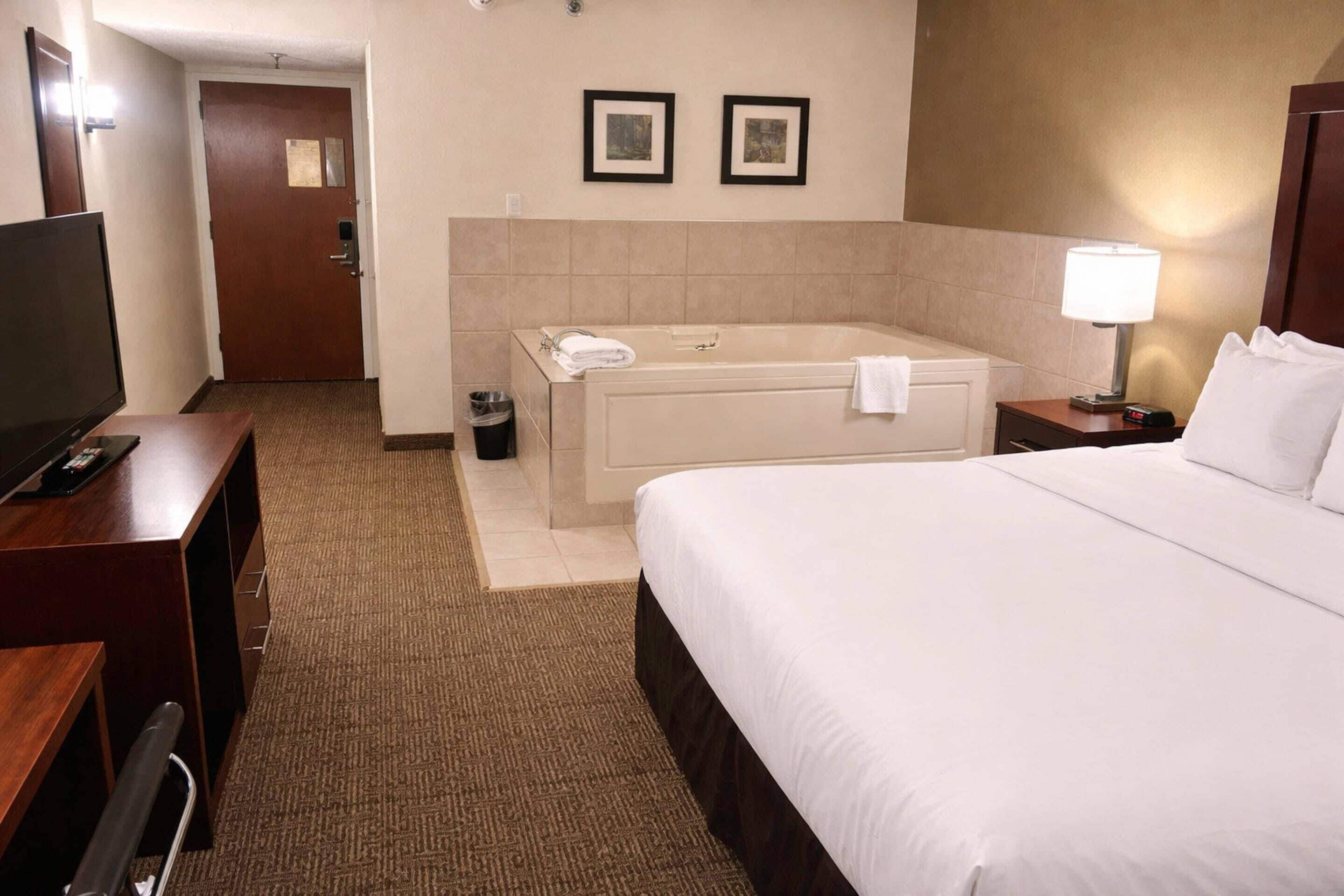 Une chambre d’hôtel avec un grand lit, une télévision, un bureau, une chaise de bureau, une salle de bain avec une baignoire et une poubelle.
