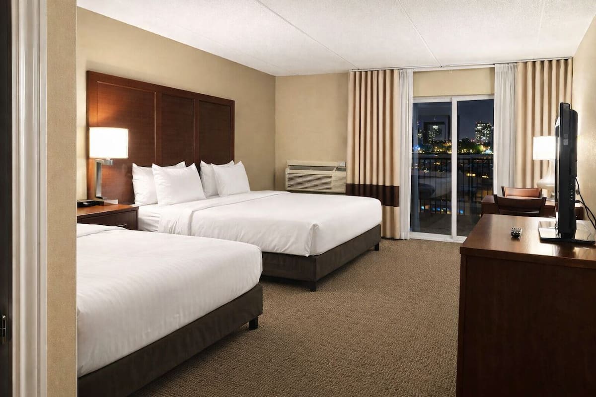 A hotel room with two beds, a desk with a TV, and a large window with curtains.