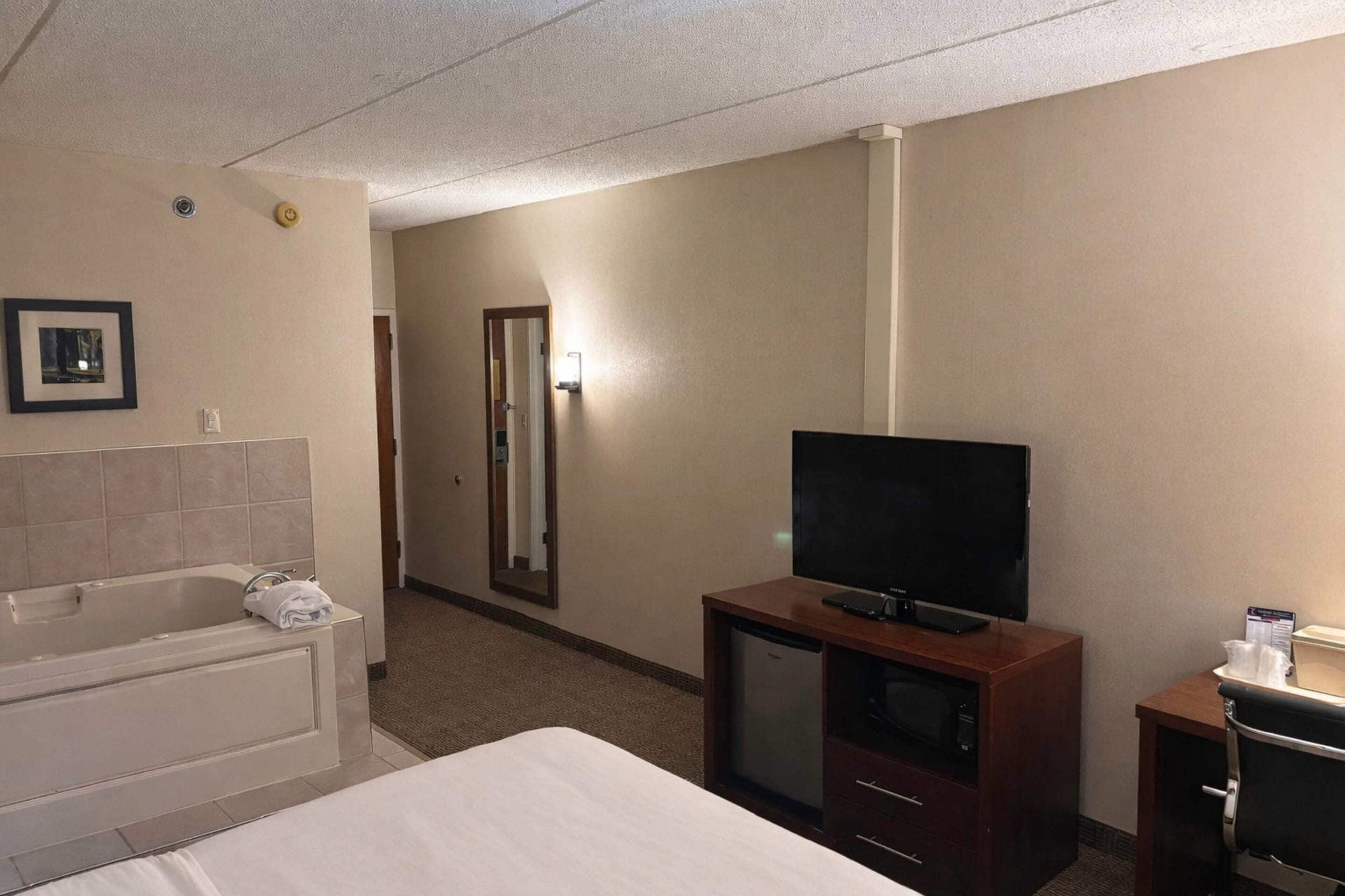 A hotel room with a bed, a TV, a small desk, a mirror, and a bathroom with a bathtub and toilet.