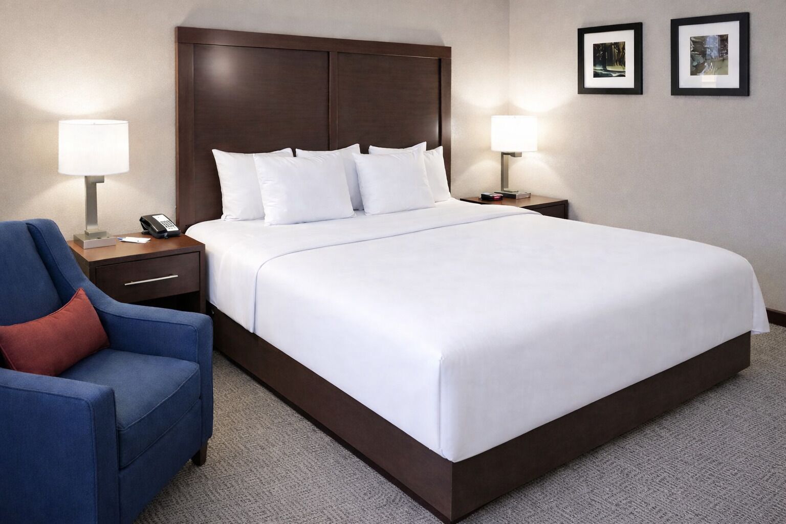 Habitación de hotel impecable con una cama grande, un sillón azul, dos mesitas de noche con lámparas y cuadros enmarcados en la pared.