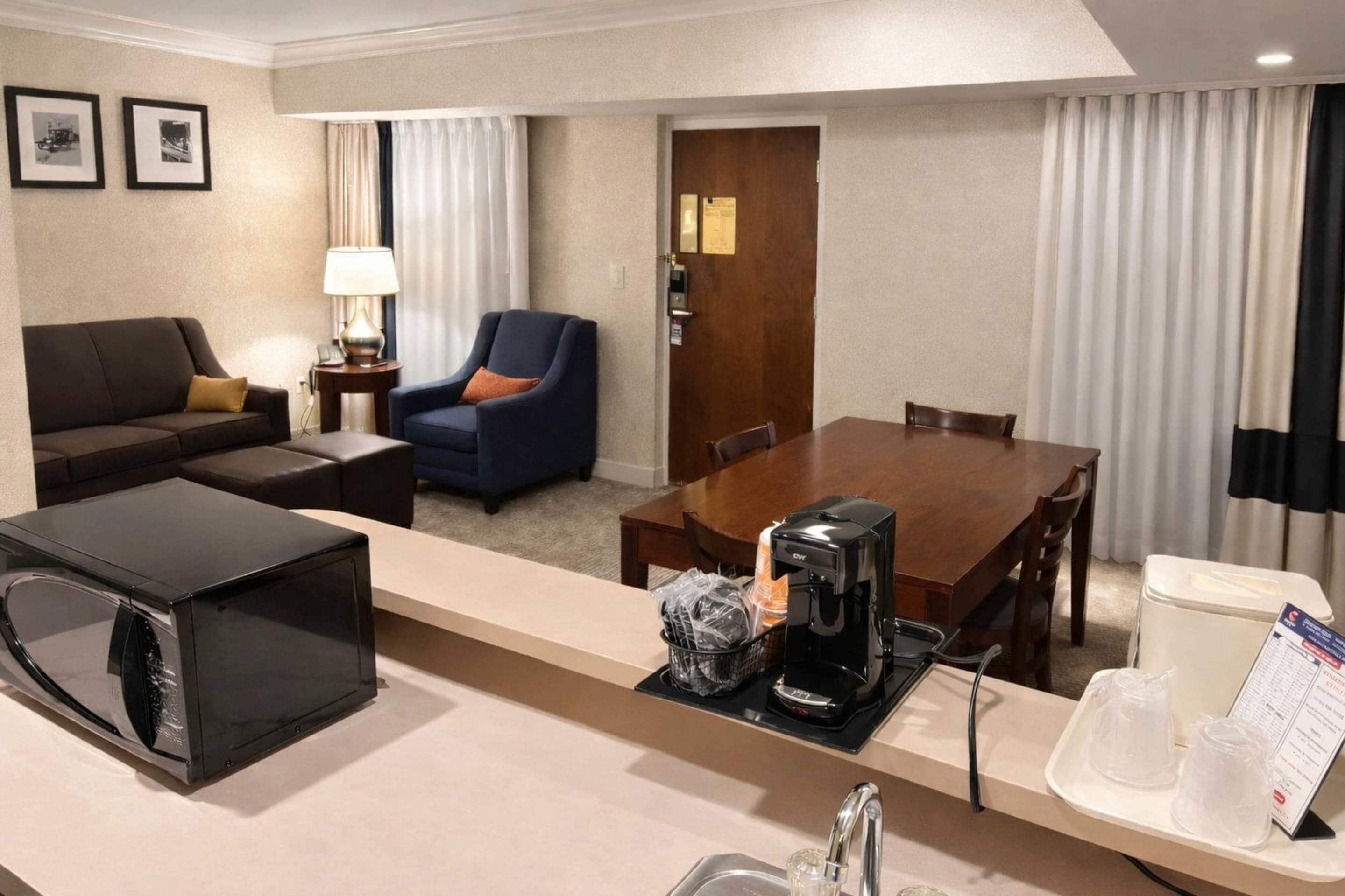 Habitación de hotel con una mesa de comedor, un sofá, una silla, microondas, cafetera, lavabo y una bandeja con jarra de café y vasos.