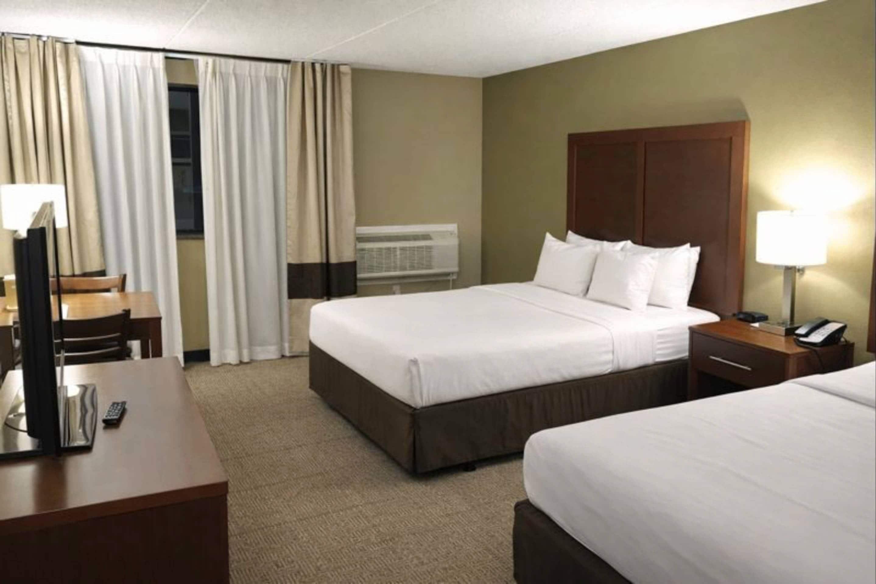 A hotel room with two beds, a desk, a chair, a lamp, and an air conditioning unit.