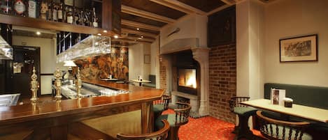 Una zona de bar con mostrador de madera, varias botellas de licores, chimenea y un área de estar con sillones tapizados en verde.