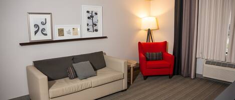 Habitación de hotel con un sofá color beige, un sillón rojo, una lámpara de pie y obras de arte en la pared.
