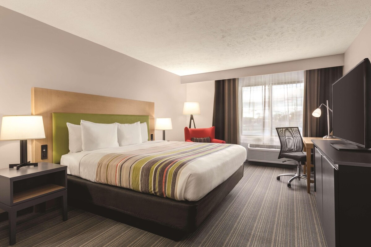 A hotel room with a large bed, two bedside tables with lamps, a desk with a chair, a TV, and a window with curtains.
