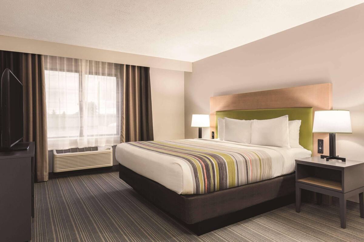 A hotel room with a large bed, two bedside tables with lamps, a flat-screen TV, and a large window with curtains.