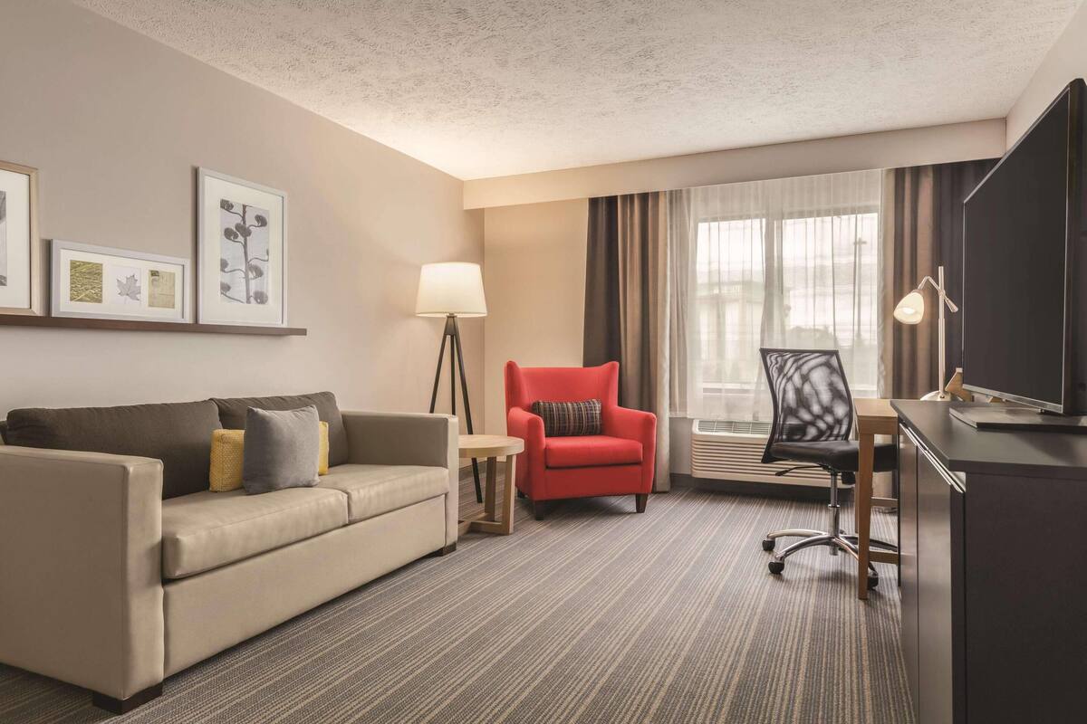 A hotel room with a sofa, a red armchair, a desk with a TV, a lamp, and framed pictures on the wall.