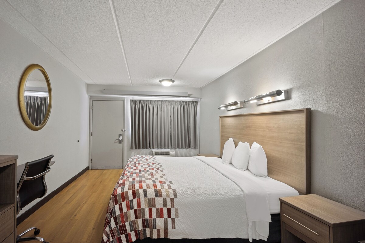 Habitación de hotel con cama, escritorio, silla, espejo y ventana con cortinas.