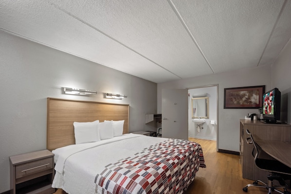 Habitación de hotel con cama, mesita de noche, escritorio, silla, televisor y baño visible a través de una puerta abierta.