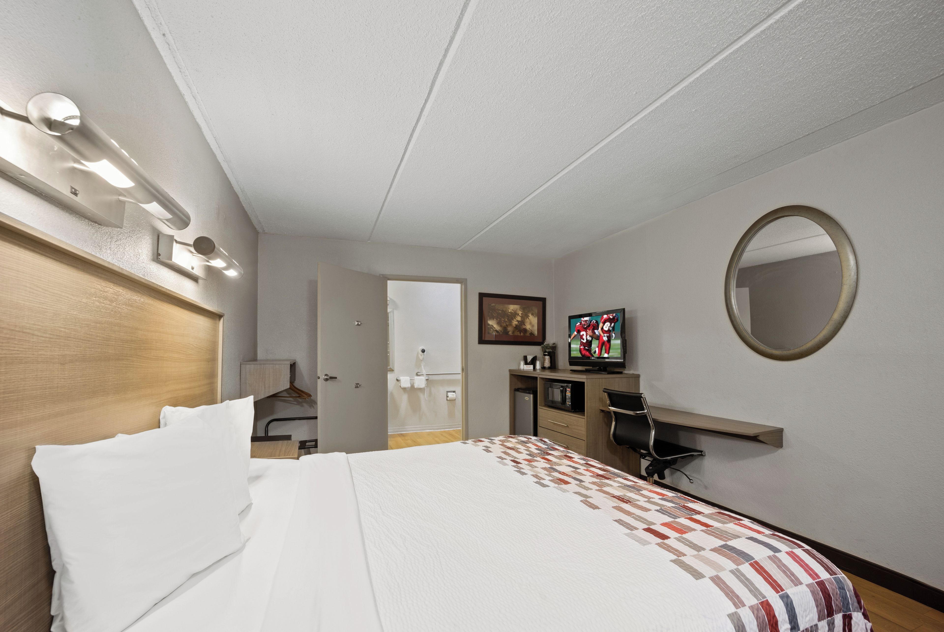 A hotel room with a bed, a television, a mirror, and a desk with a chair.