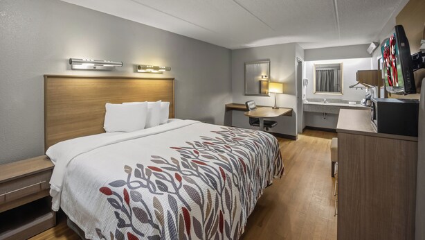 A hotel room with a bed, bedside tables, a desk, a chair, a mirror, a television, and a bathroom with a sink and mirror. - Red Roof Inn Greensboro Airport (Greensboro)