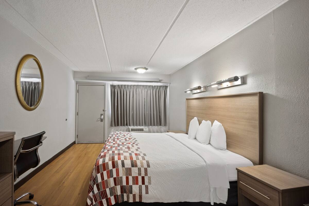 Habitació d'hotel amb un capçal de llit de fusta, un llit gran amb roba de llit blanca, un escriptori amb una cadira, un mirall i una finestra amb cortines.