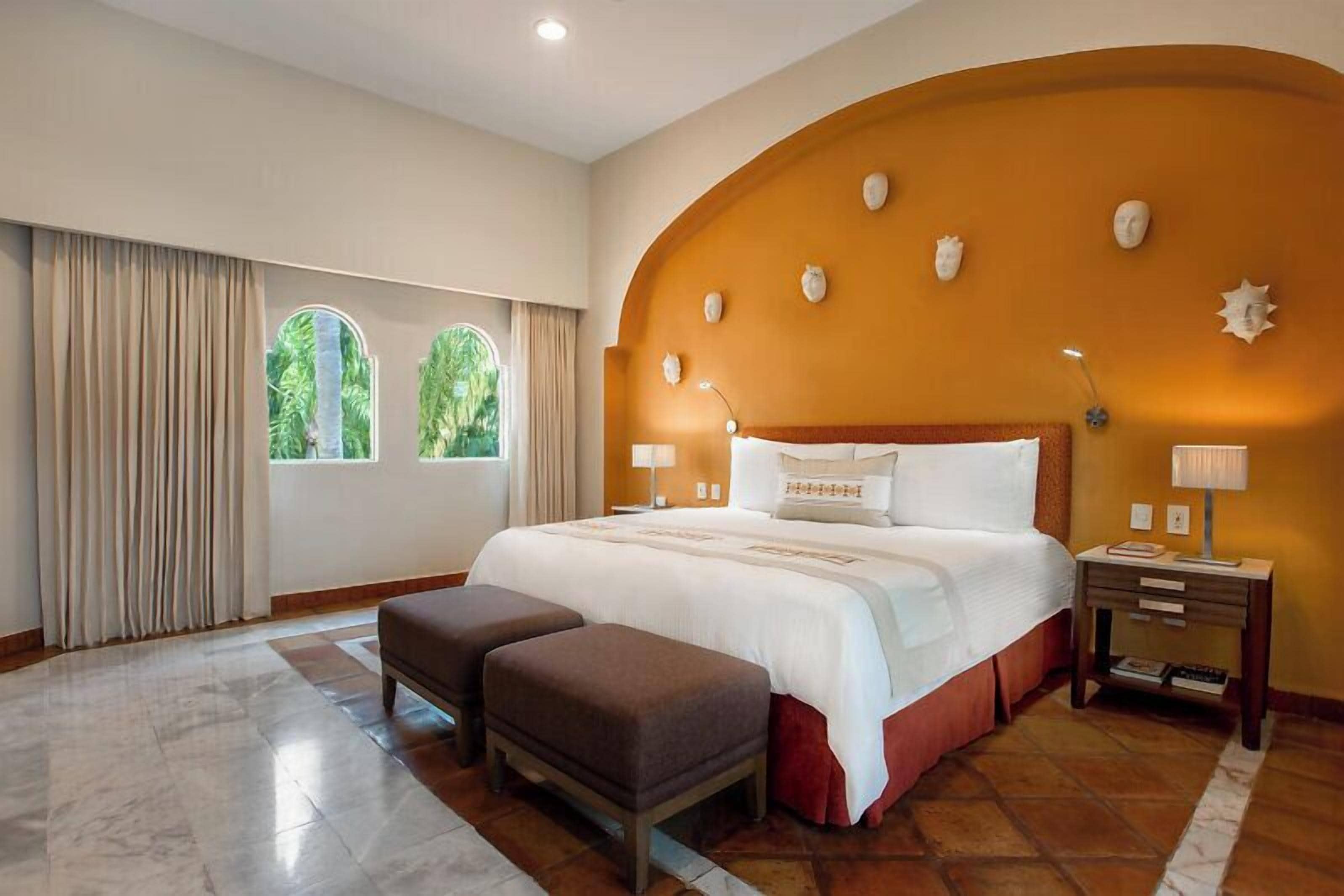 Una habitación de hotel con una cama grande, dos mesitas de noche, una ventana con cortinas y una pared con detalles decorativos.