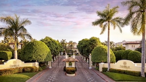 A gated entrance with a sign reading "CASAVELAS" surrounded by well-maintained hedges and palm trees.