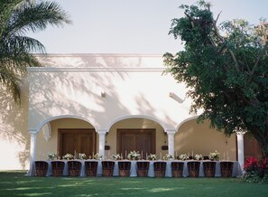 An outdoor dining area with a long table set for a meal, flanked by three wooden doors and a white building facade.
