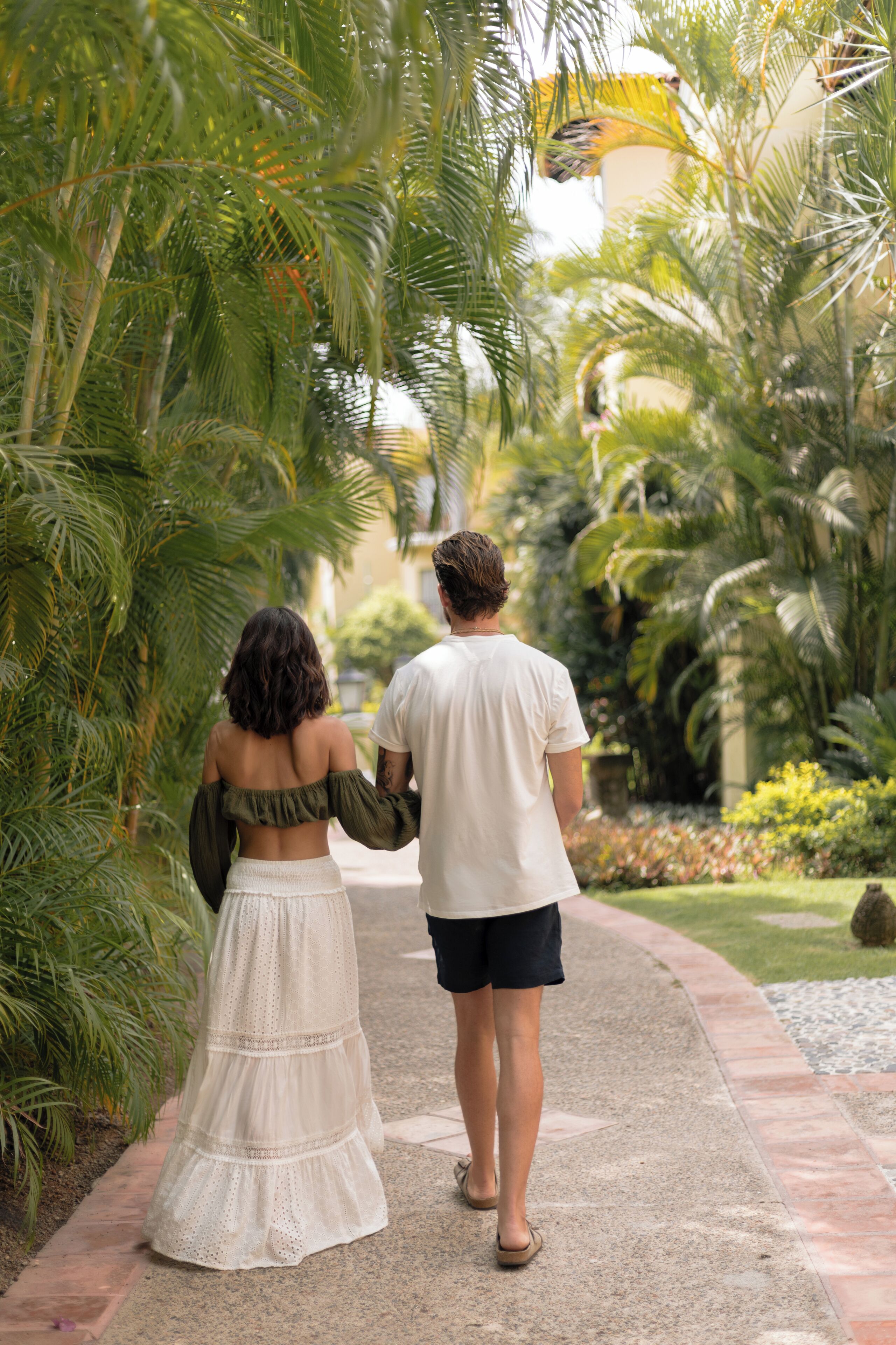 A couple walking on a paved path surrounded by lush greenery and palm trees.
