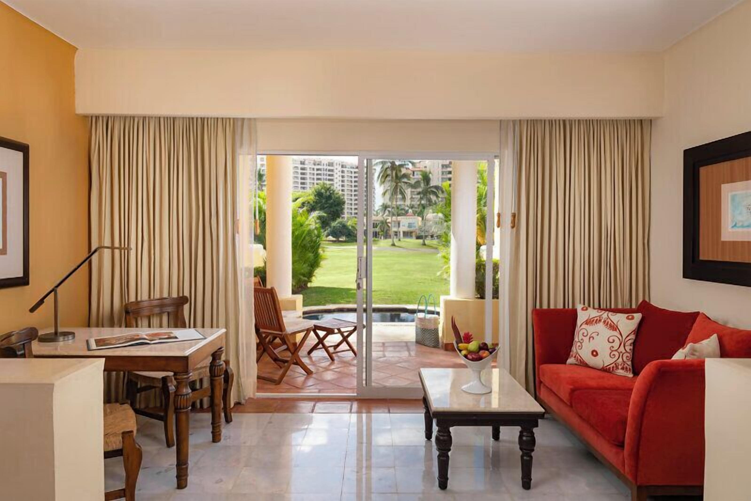 Habitación de hotel con comedor, sofá rojo, mesa de centro y vistas a un jardín y edificios.