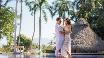 A couple standing by a pool with palm trees and a thatched-roof hut in the background.