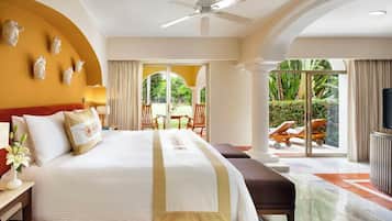 A hotel room with a large bed, a ceiling fan, a view of the outdoor garden, and a seating area with chairs and a table.