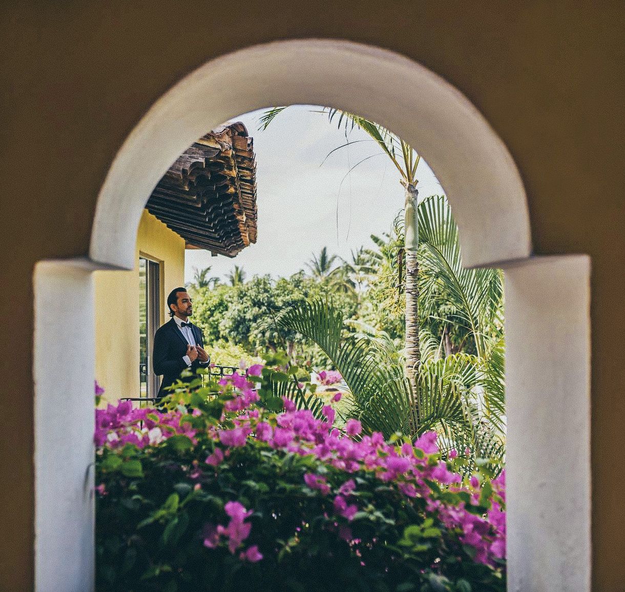 A man in a suit standing at an archway with a view of a yellow building and lush greenery.