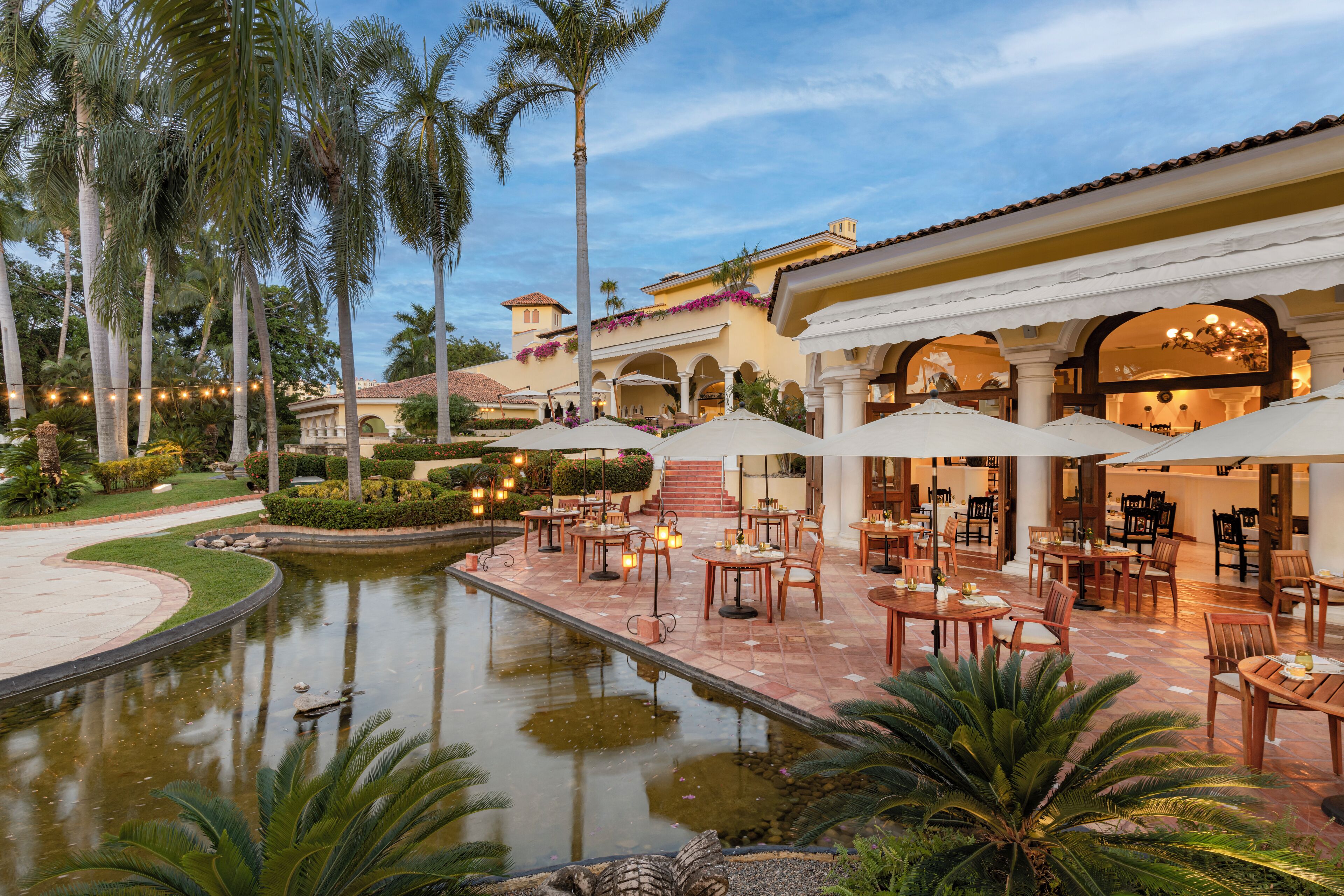 Un patio con piscina, mobiliario exterior, palmeras y un edificio con arcos y columnas.