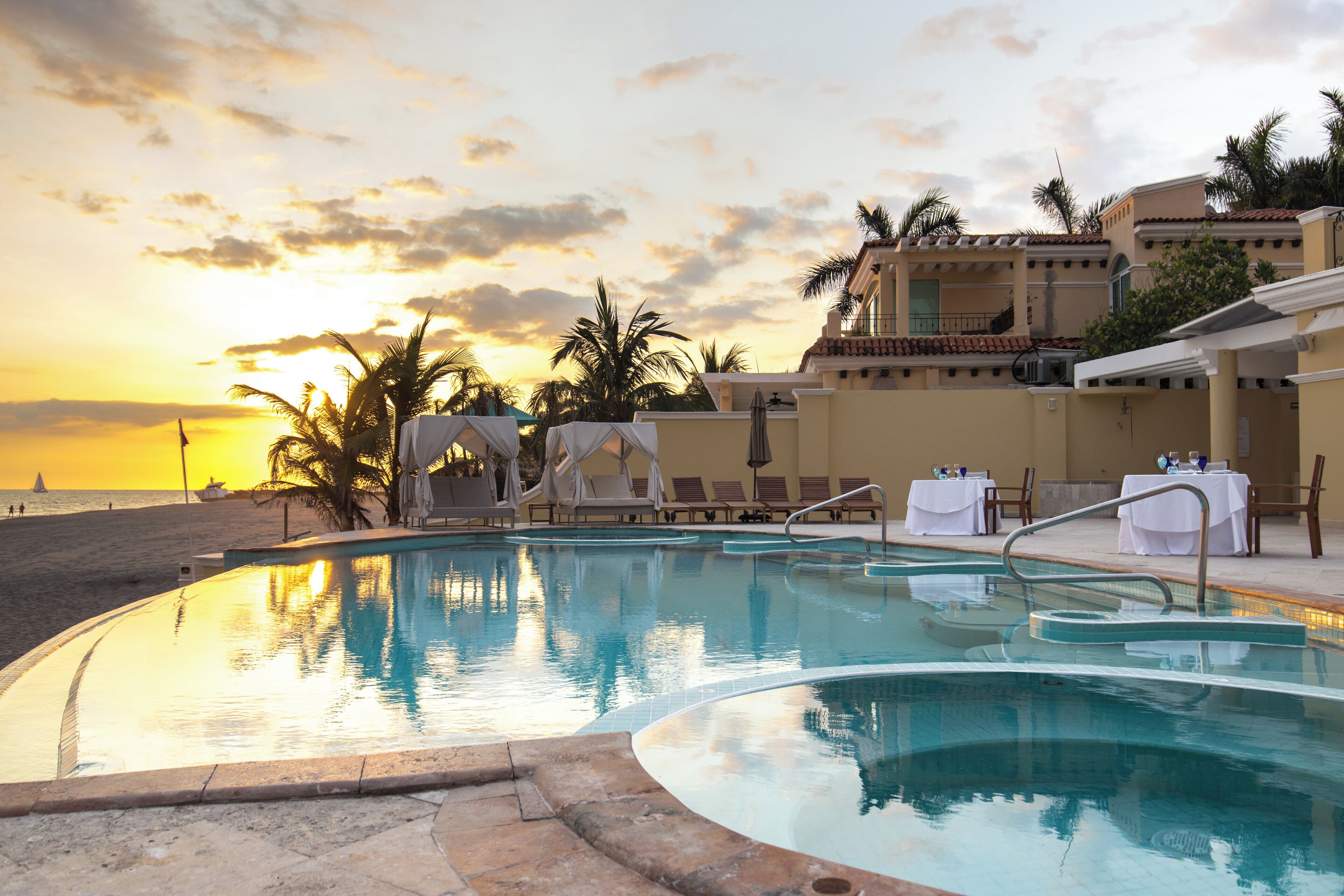 A pool area with lounge chairs and tables, a sunset view, and a building with a tiled roof.