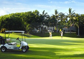 A golf course with a golf cart, palm trees, and a building in the background.