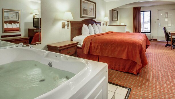 A hotel room with a large bed, a sitting area with a chair, a desk, a television, a ceiling fan, and a bathtub.