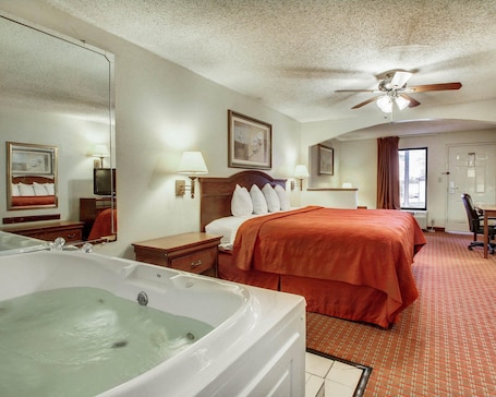 A hotel room with a large bed, a sitting area with a chair, a desk, a television, a ceiling fan, and a bathtub.