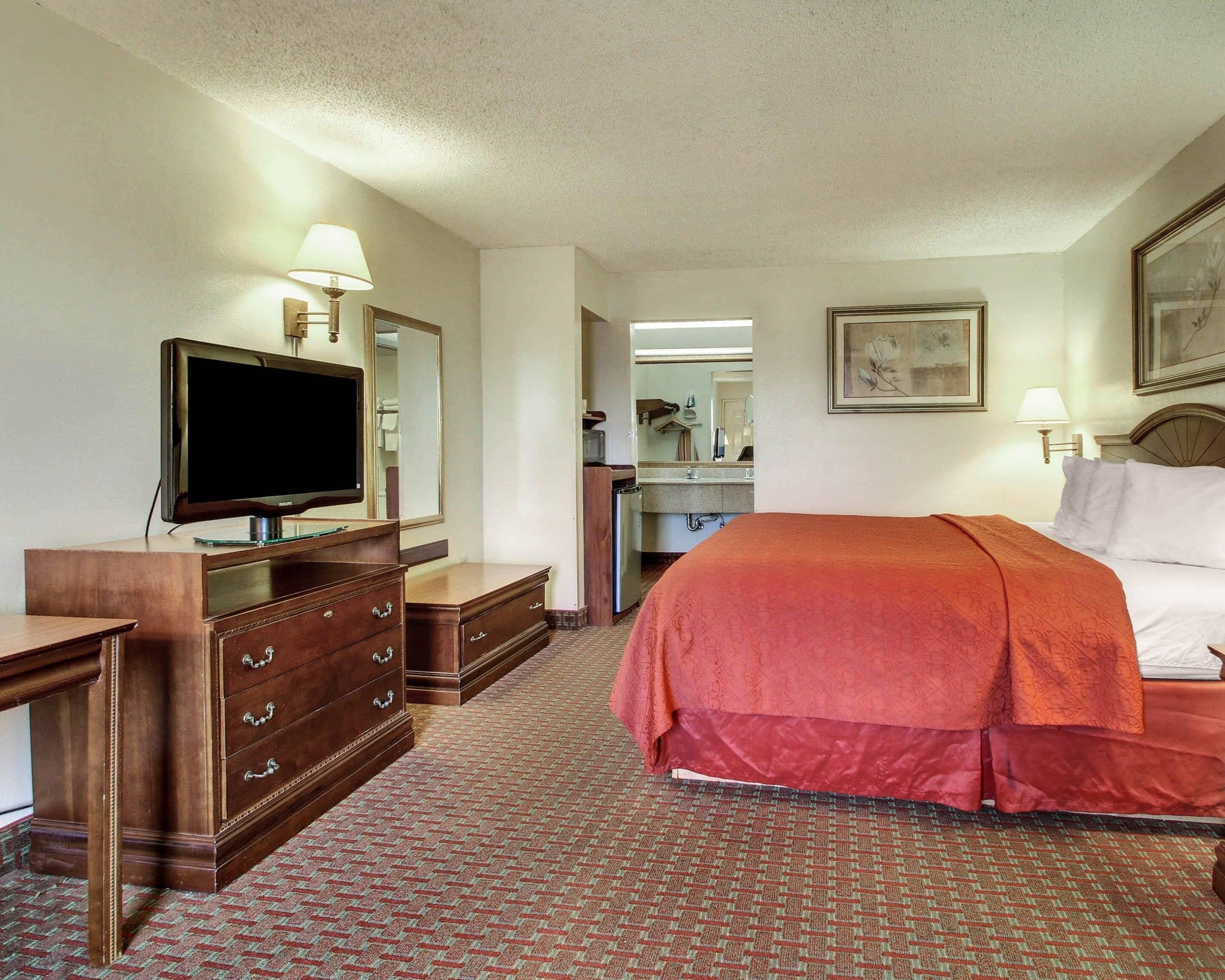Une chambre d’hôtel avec un lit, une télévision, une commode, un miroir et une salle de bain visible à travers une porte ouverte.