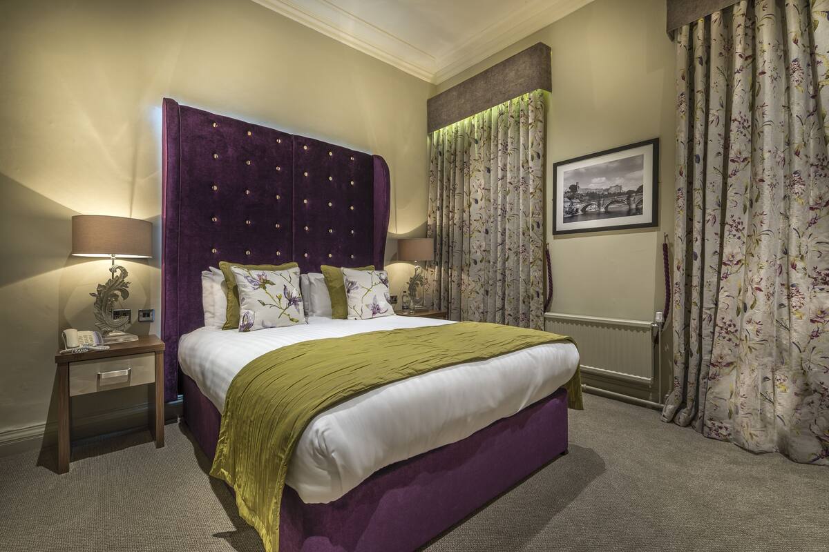 A bedroom with a large bed, two bedside tables, a nightstand, a lamp, a framed picture on the wall, and a window with curtains.