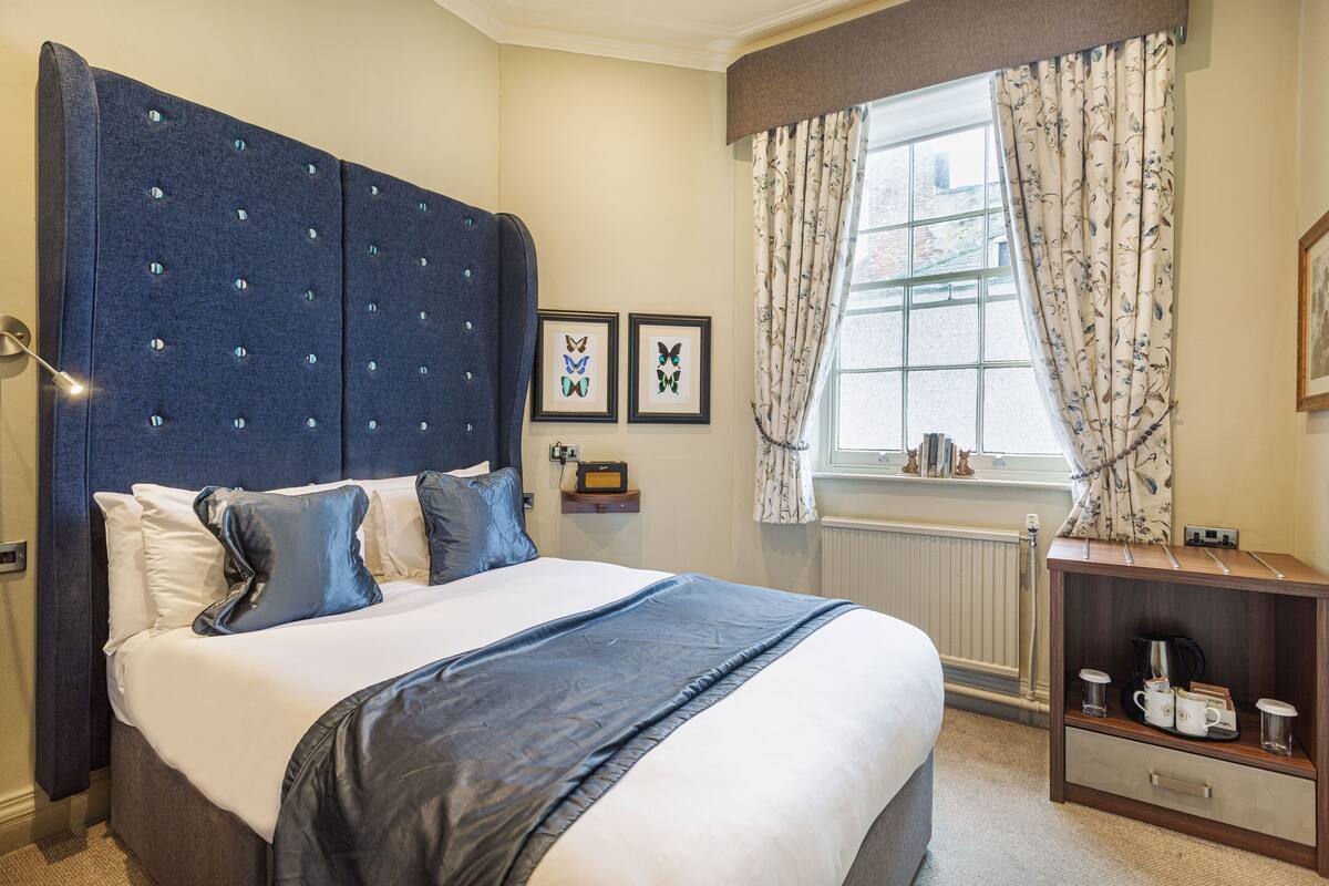 A bedroom with a large bed, a headboard, a bedside table with a radio, and a window with curtains.