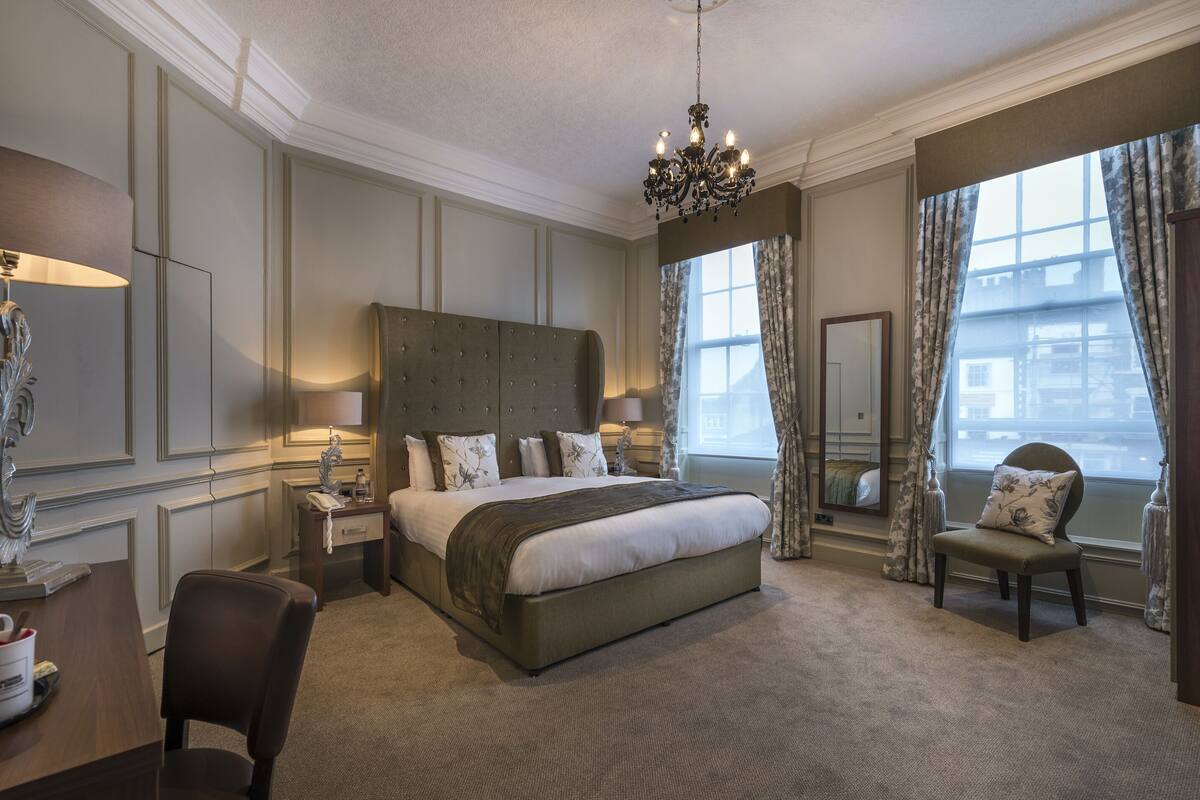 A spacious bedroom with a large bed, a desk, and a chair. There is a chandelier hanging from the ceiling and a window with curtains.