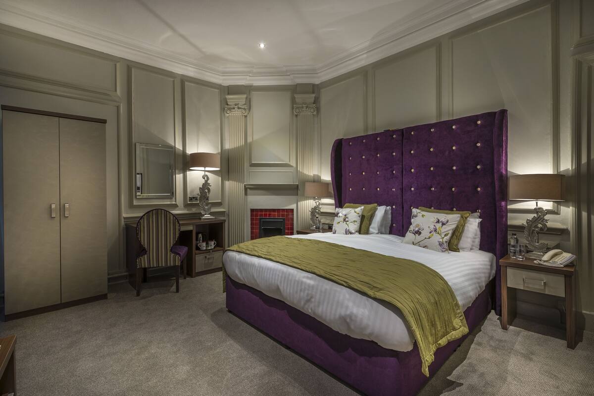 A bedroom with a large bed, a desk, a chair, a wardrobe, and a nightstand with a lamp.