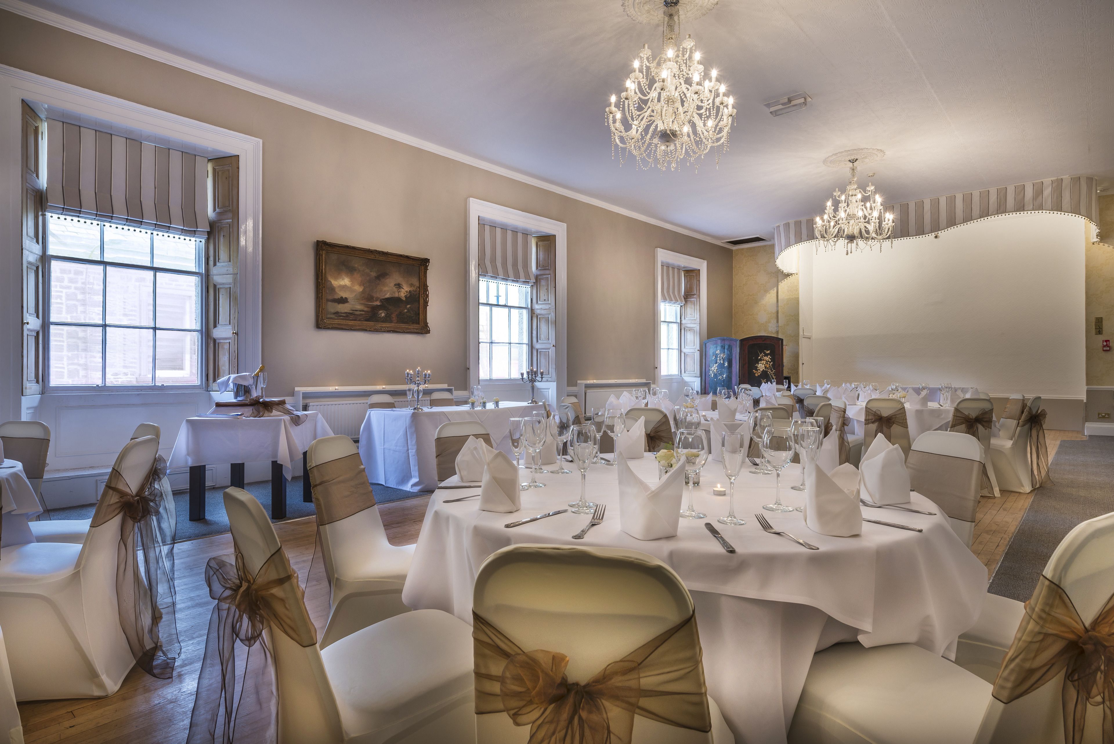 A banquet hall with round tables set for a formal event, featuring white tablecloths and chairs with gold sashes.