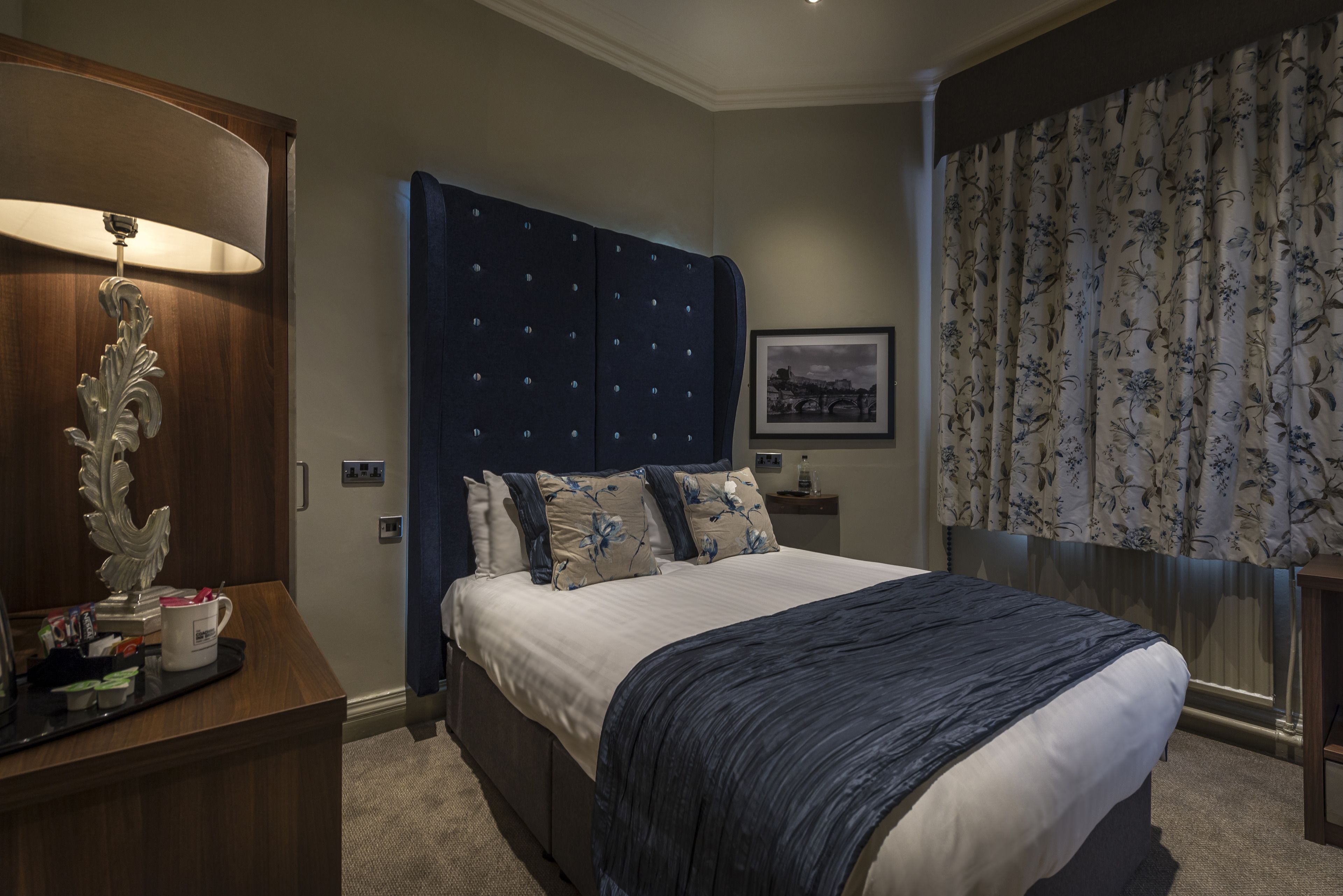 A neatly arranged bedroom with a large bed, a bedside table with a lamp, and a framed picture on the wall.