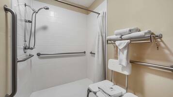 Une salle de bain avec des murs recouverts de carreaux blancs, une douche équipée d’une barre d’appui, un porte-serviettes et des toilettes blanches.