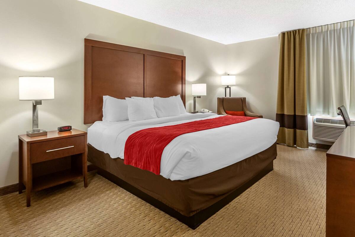 A hotel room with a large bed, two bedside tables with lamps, a chair, and an air conditioning unit.