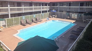 A swimming pool with lounge chairs and umbrellas, surrounded by a fenced area with a multi-story building in the background.