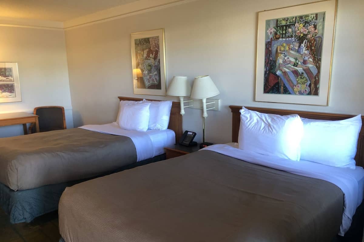 A hotel room with two beds, a desk, a chair, and two paintings on the walls.