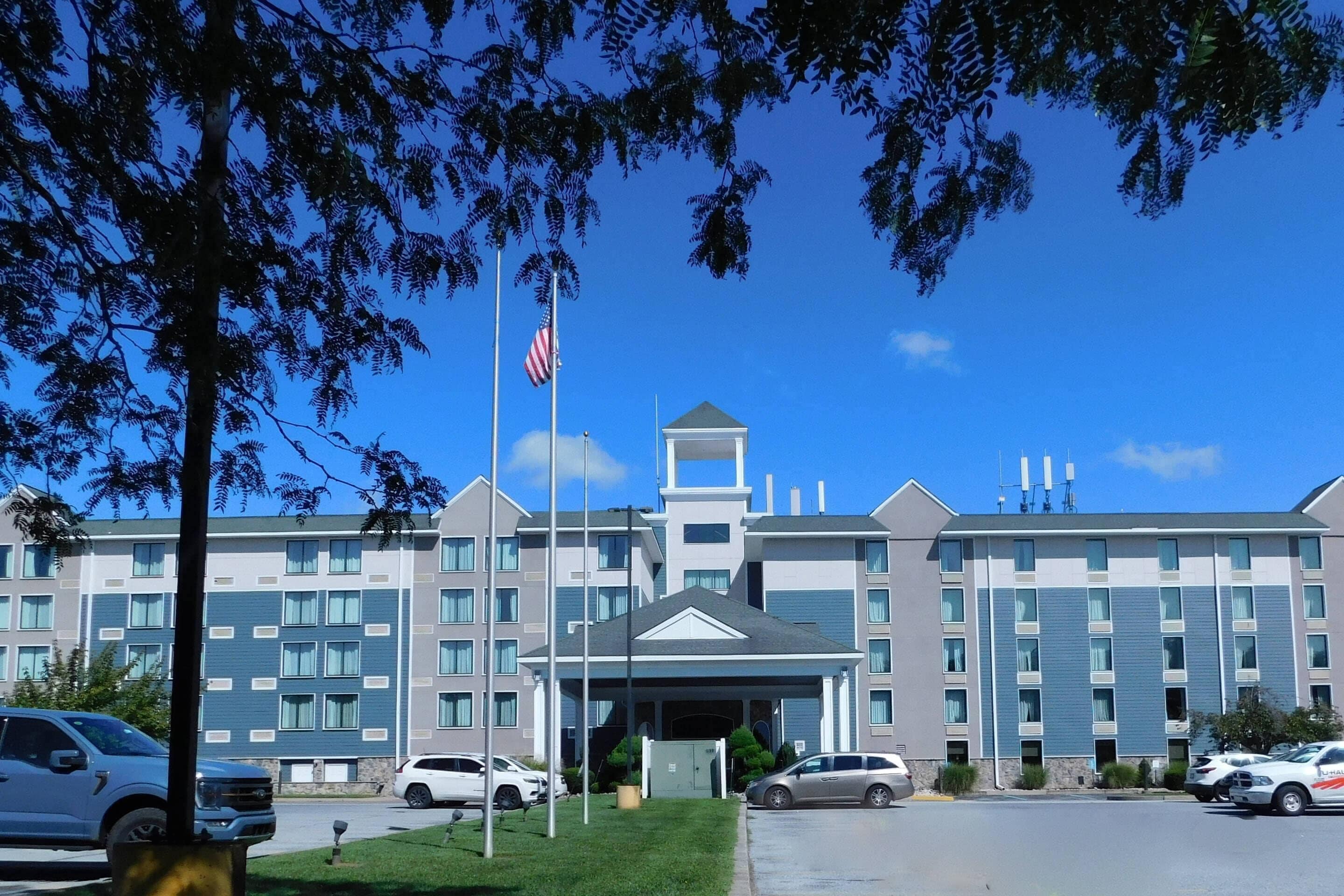 Un hotel de varios pisos con entrada central, bandera y zona de estacionamiento.