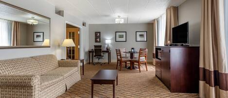 A hotel room with a sofa, a dining table with chairs, a TV, a desk with a chair, and framed pictures on the wall.