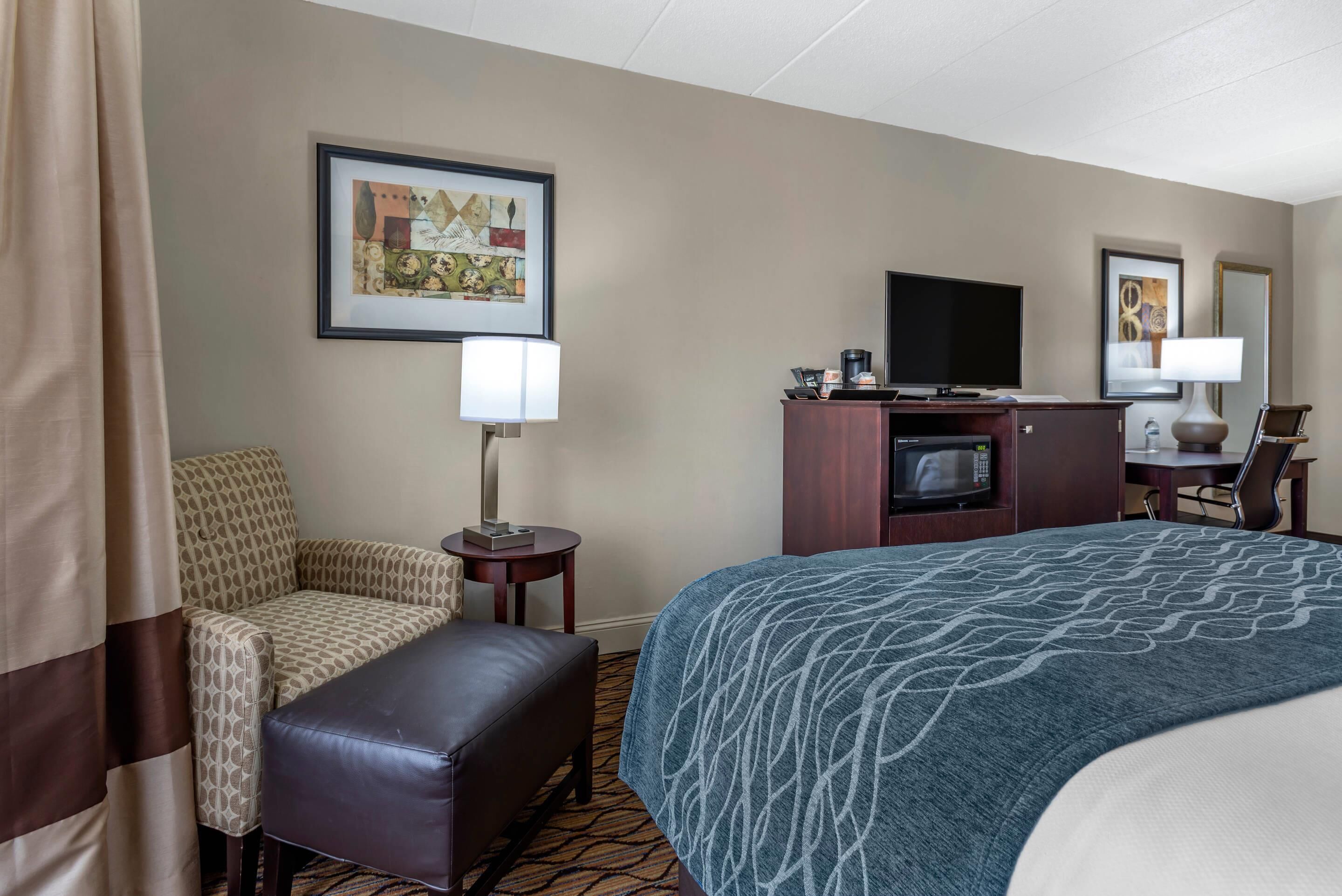 A hotel room with a bed, a sofa, a chair, a small table with a lamp, a TV, and a microwave.