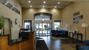 A reception area with a wooden desk, a blue sofa, and a small table with a lamp. There is a large window with a view of the outside. - Comfort Inn & Suites Glen Mills - West Chester (Glen Mills)