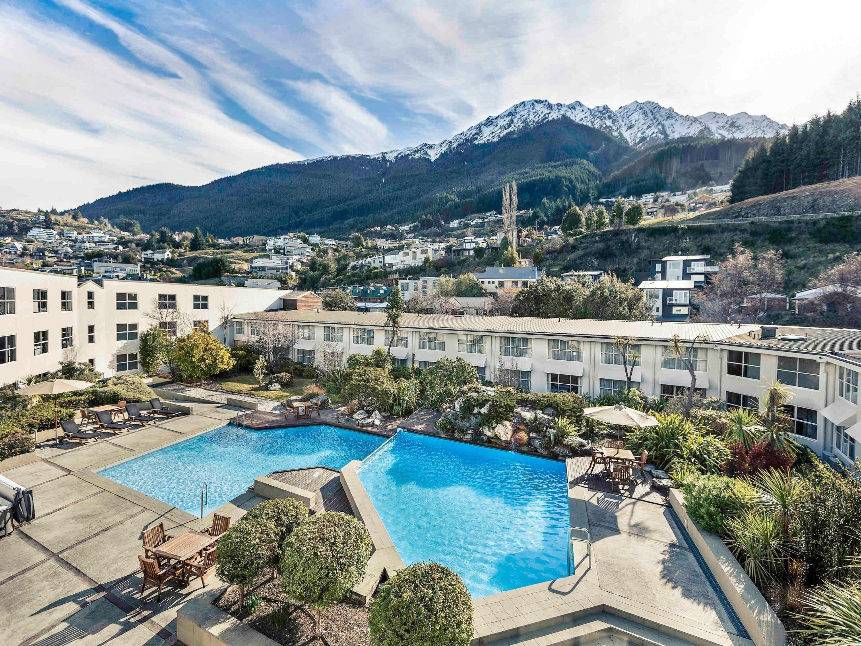 A hotel with two swimming pools, a patio area, and a view of snow-capped mountains.