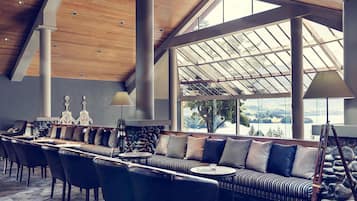 Un amplio salón con techo alto, ventanas grandes que ofrecen vistas al exterior, un sofá largo con numerosos cojines y una pared de piedra.