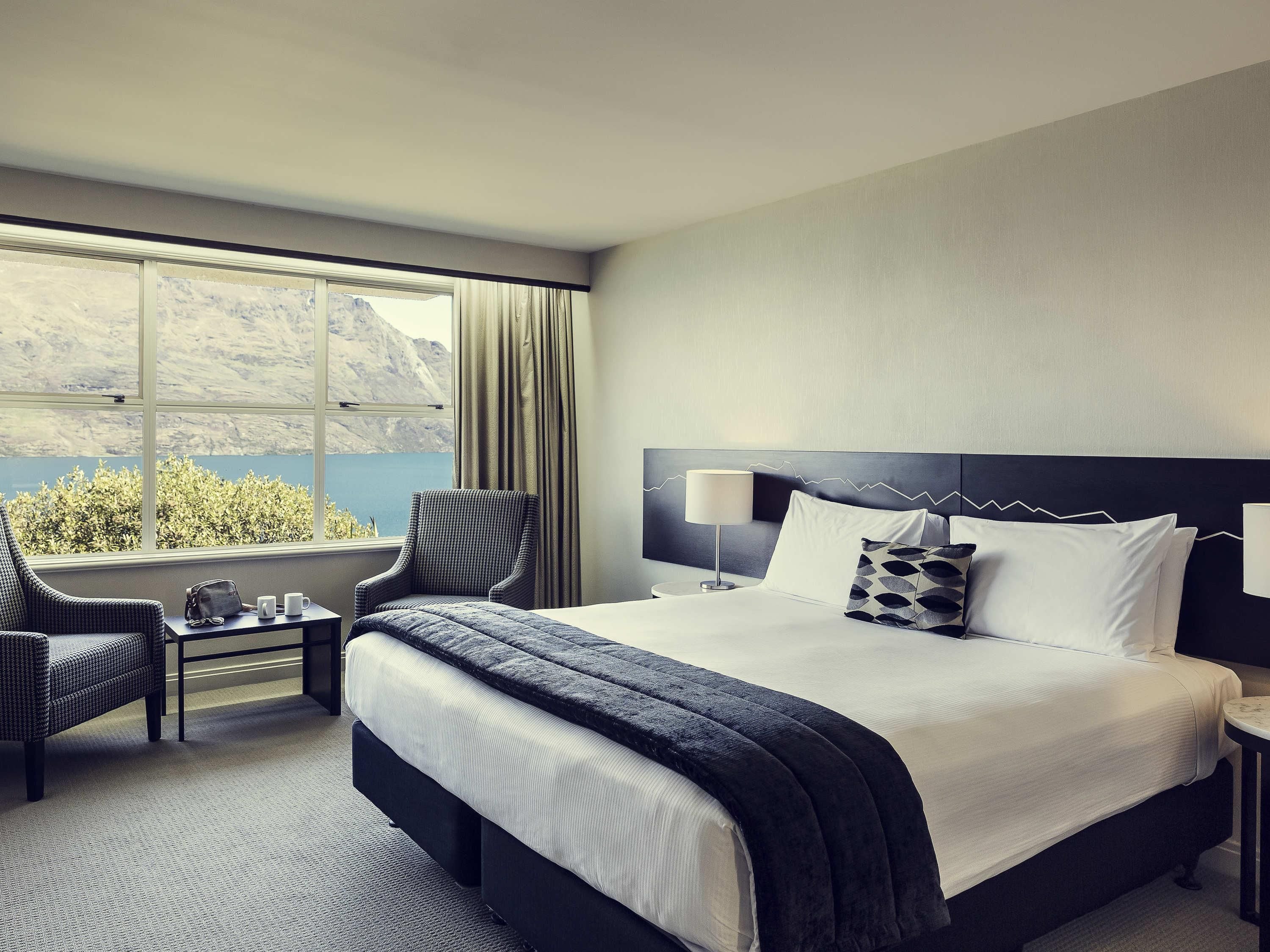 A hotel room with a large bed, a chair, a small table, and a view of the mountains and lake.