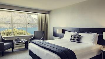 A hotel room with a large bed, a chair, a small table, and a view of the mountains and lake.