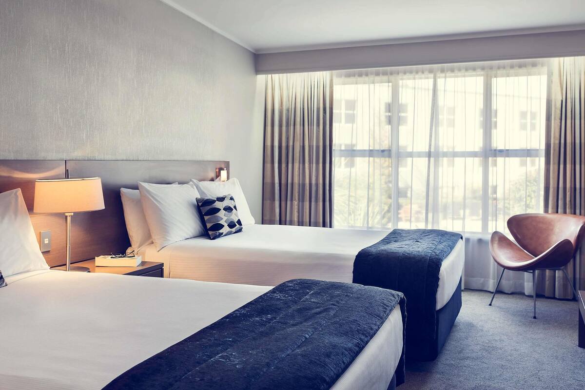 Ein Hotelzimmer mit zwei Betten, einem Sessel, einer Lampe und einem Fenster mit Vorhängen.