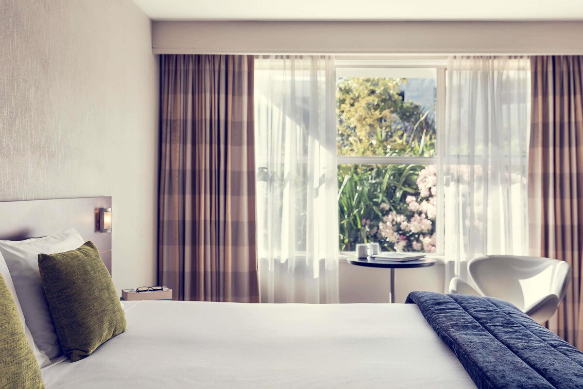 A hotel room with a bed, a small table, a chair, and a window with curtains.
